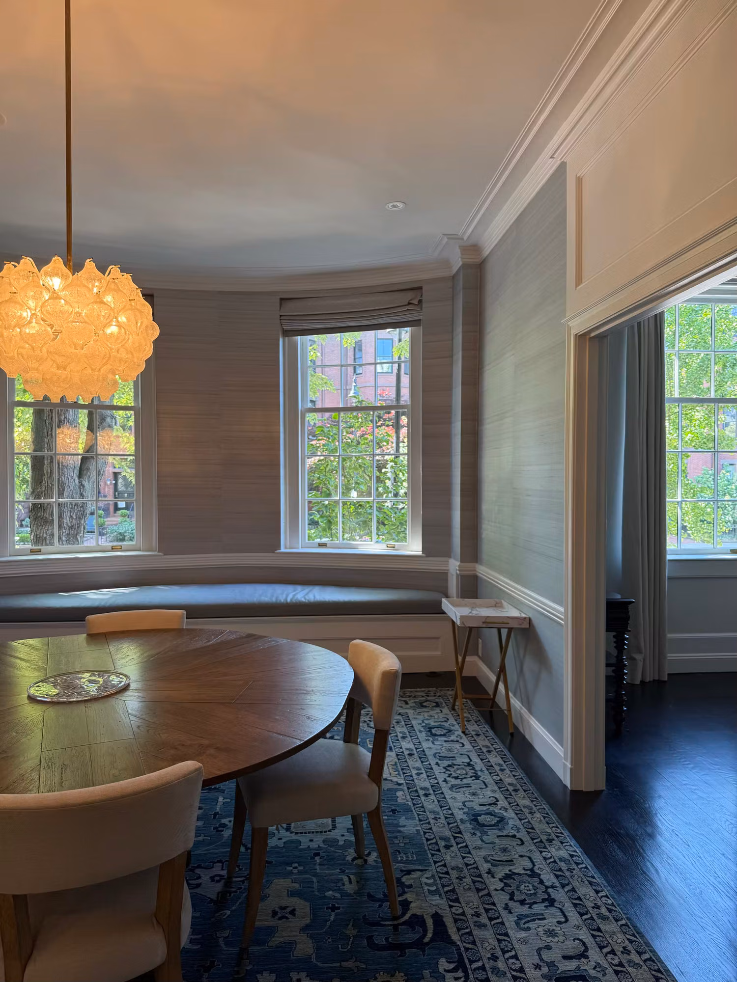 Dining room with a curved bay of clear divided-light windows, round wood table, and grasscloth-clad walls.