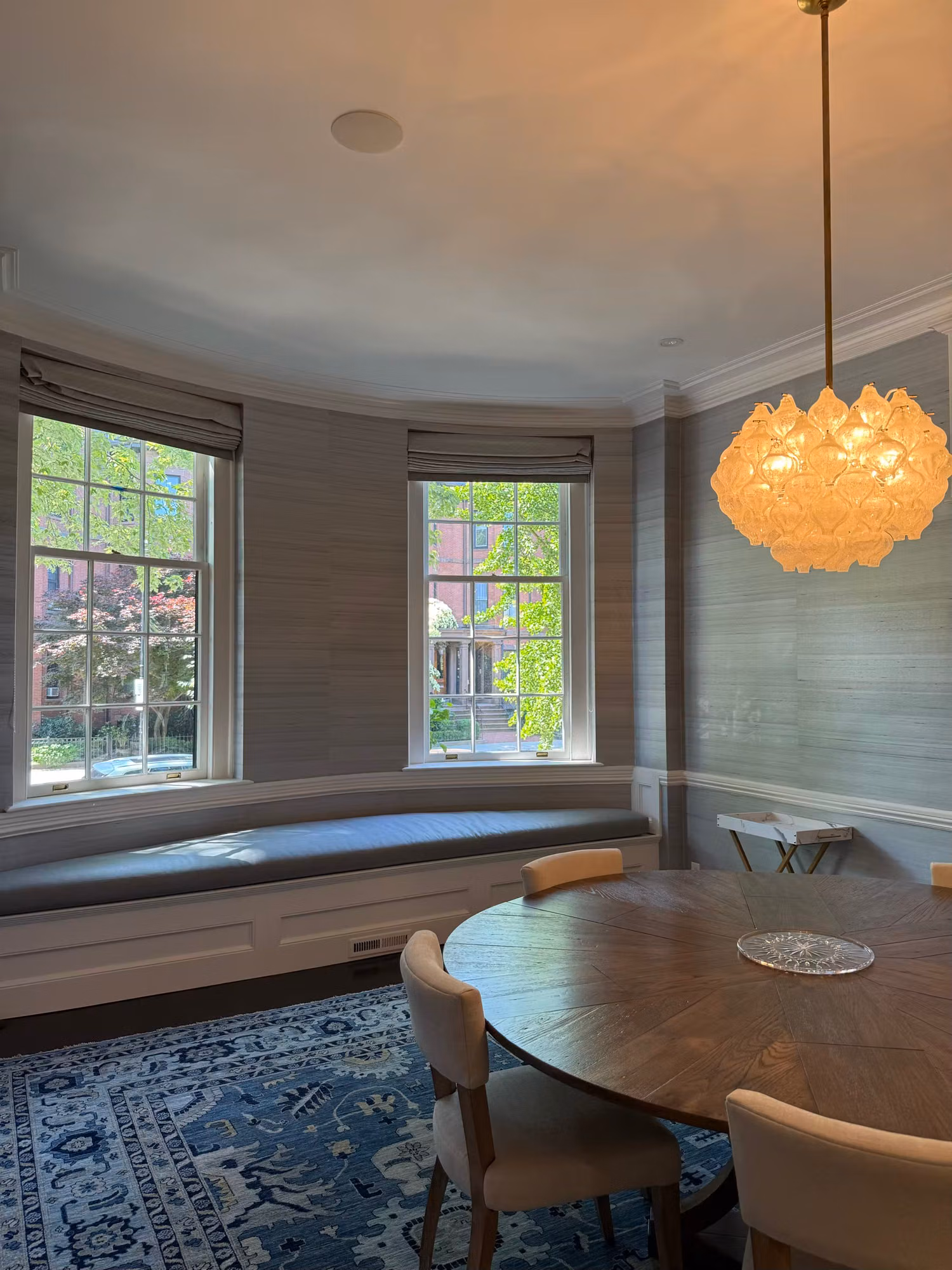 Dining room bay of clear divided-light windows with shades and a window seat beside a round table and pendant light.