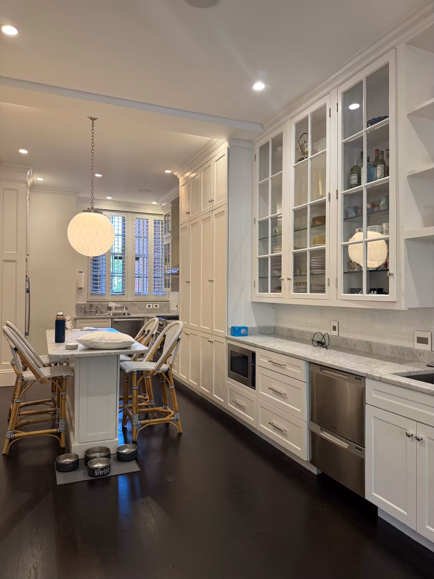 Bright white kitchen with glass-front cabinets and a shuttered window above the sink letting in daylight.