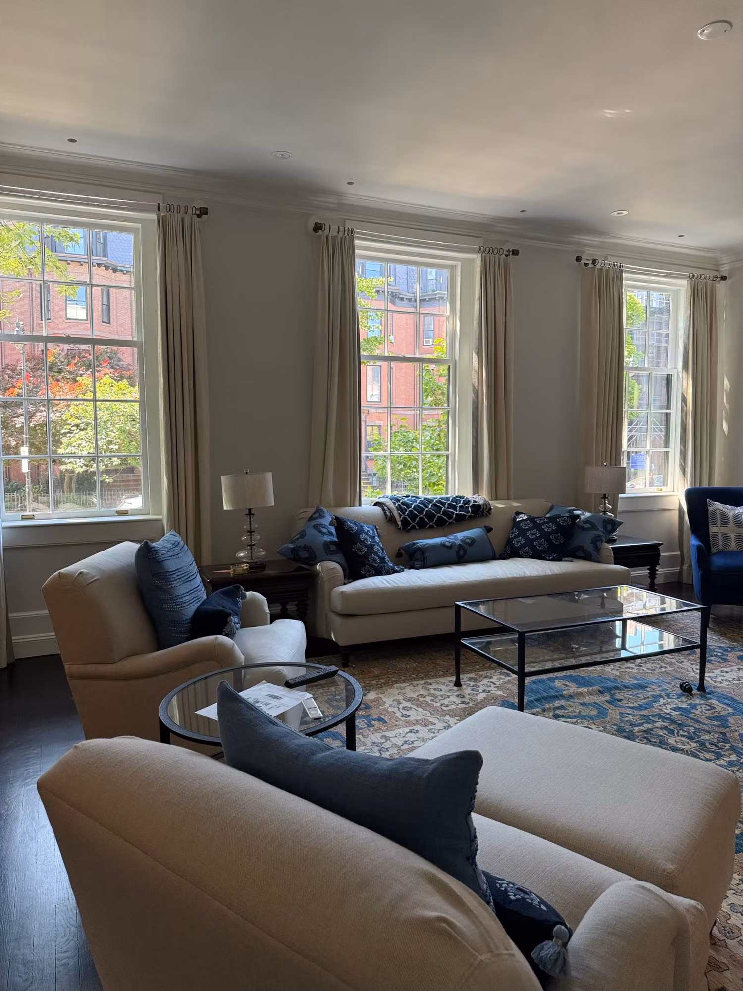 Furnished living room with several tall divided-light windows dressed in long drapes overlooking a street.