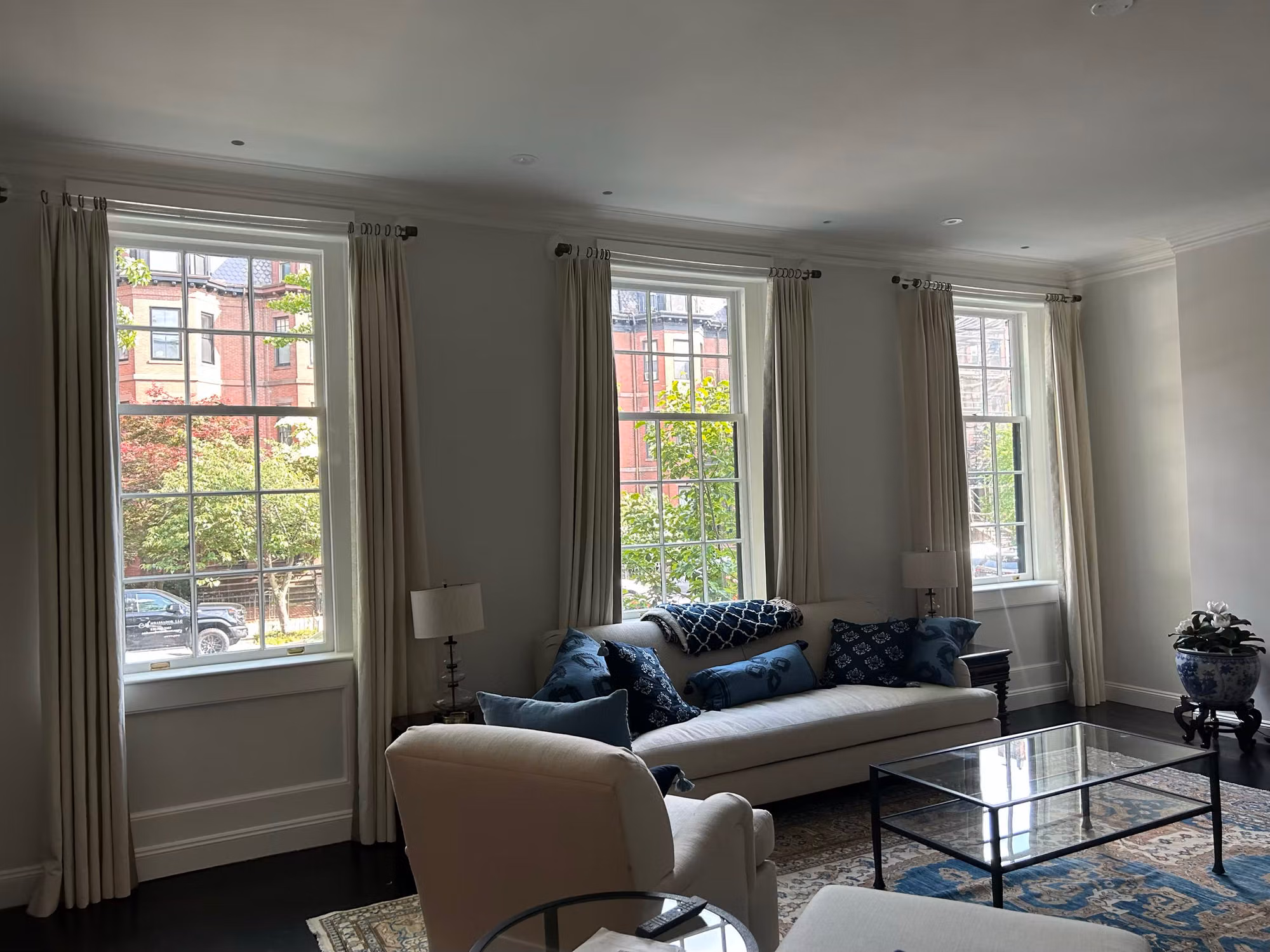 Living room with three tall double-hung grid windows framed by neutral drapes and a sofa with city views beyond.