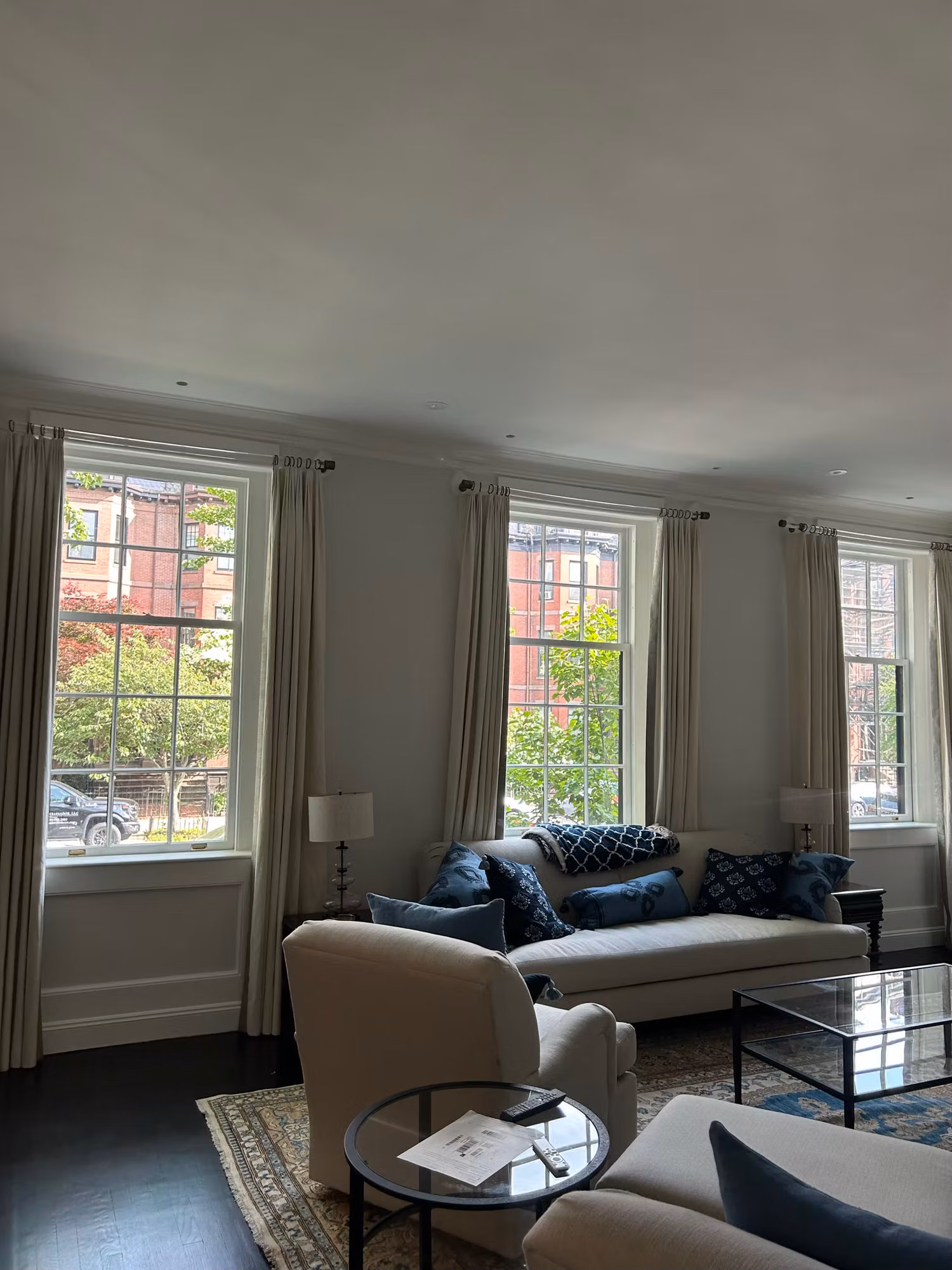 Bright living room with tall grid windows and drapes, blue accent pillows on a sofa, and brick rowhouses visible outside.