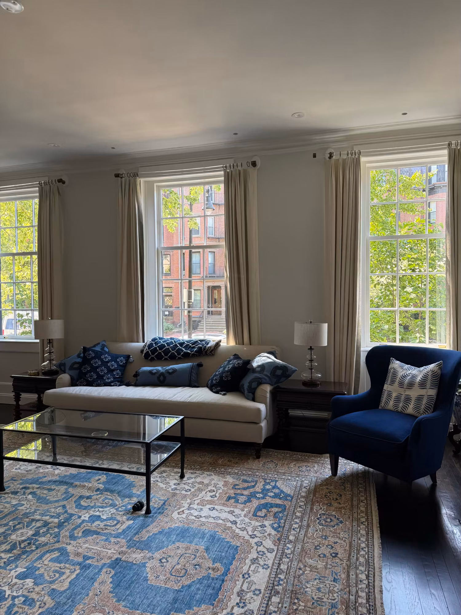 Living room seating area in front of two large divided-light windows with floor-length drapes and a navy armchair.