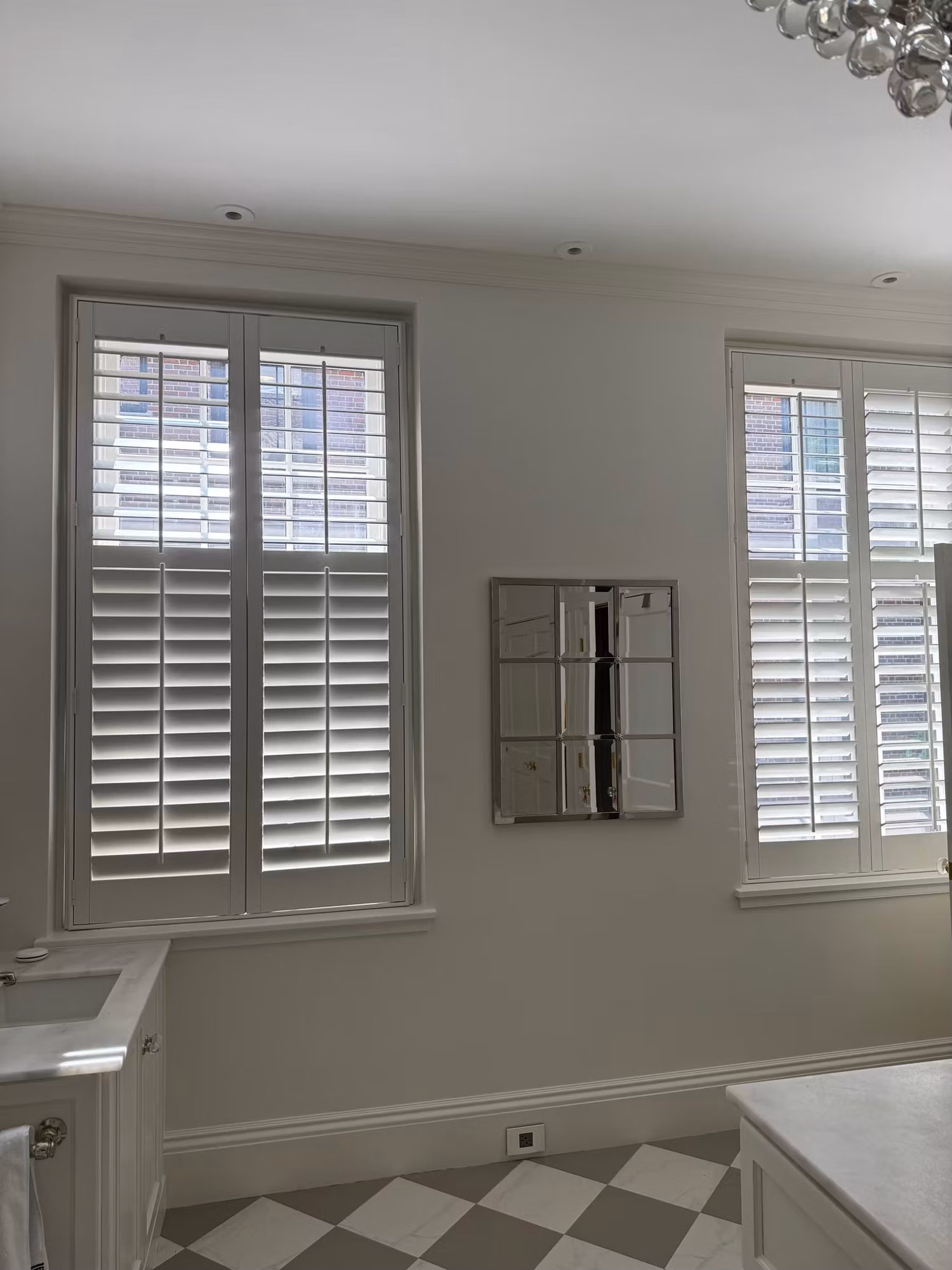 Two residential bathroom windows fitted with white plantation shutters above a checkered tile floor.