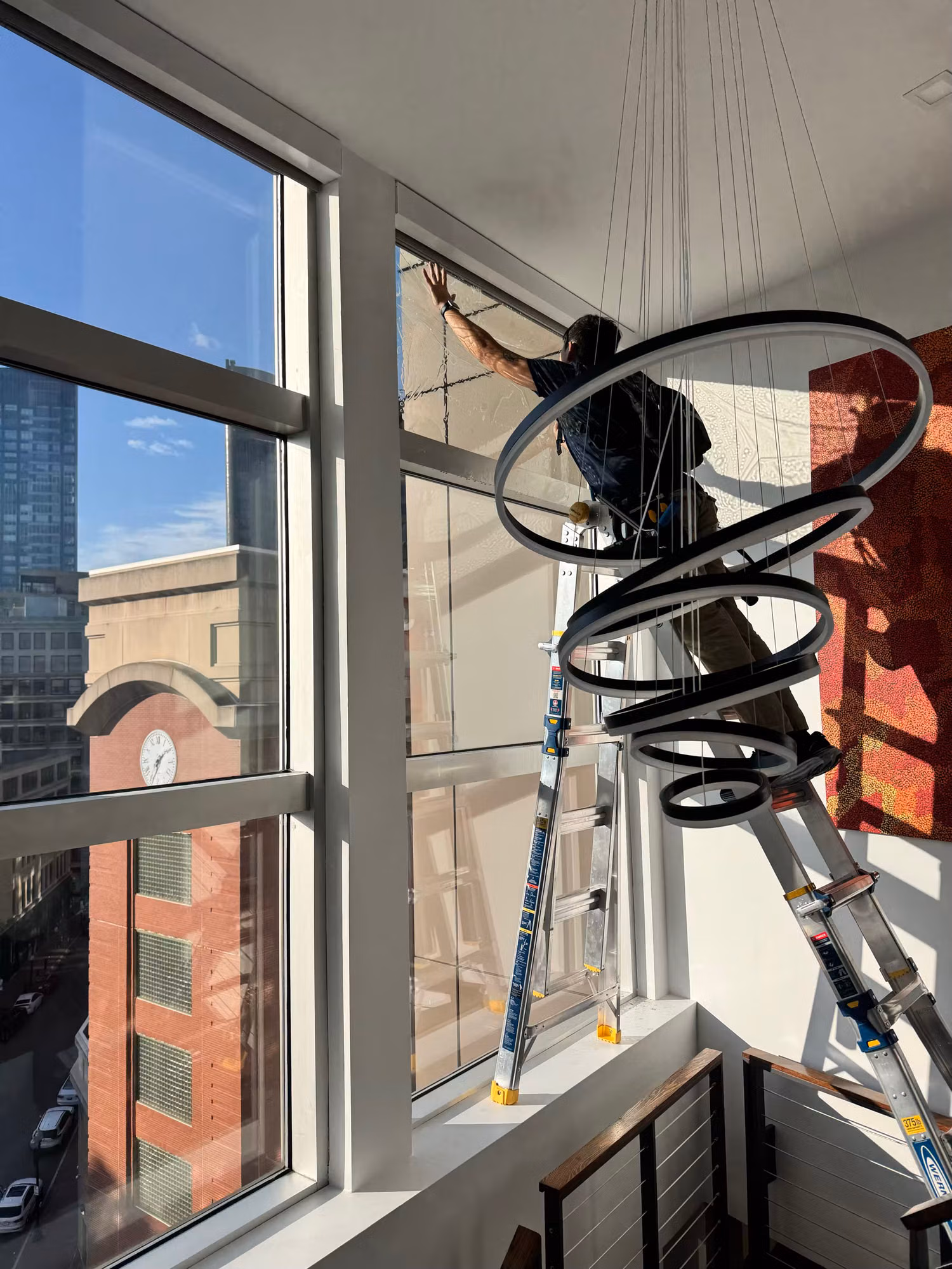 Worker reaching across tall stairwell windows to install film beside a spiral ring light fixture.