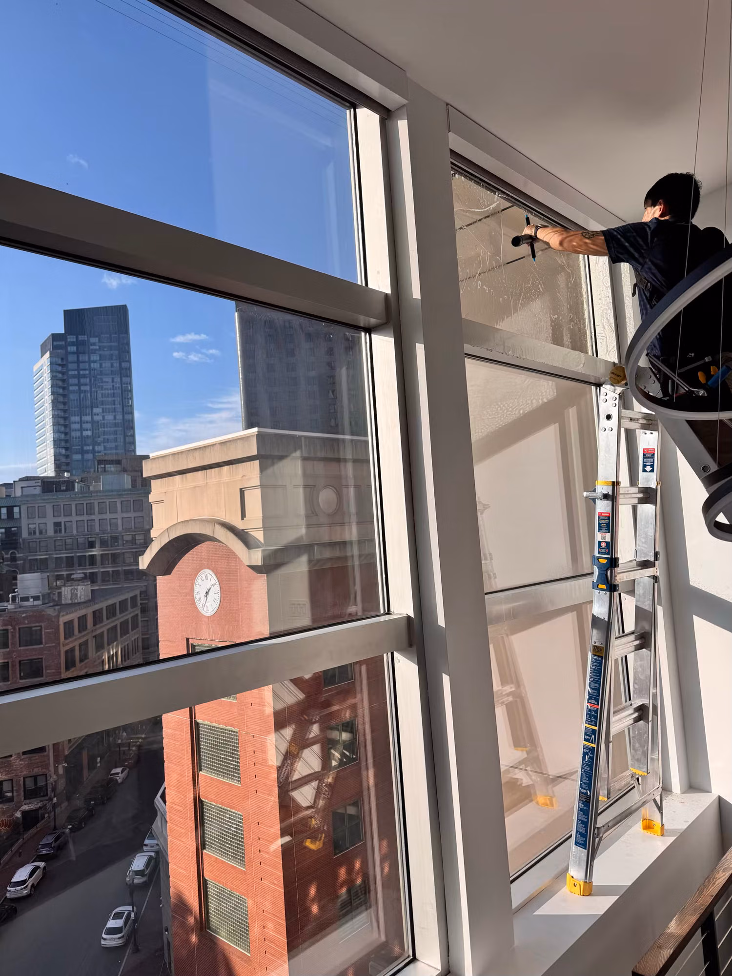 Technician installing film on large floor-to-ceiling penthouse windows with a city skyline view beyond the glass.