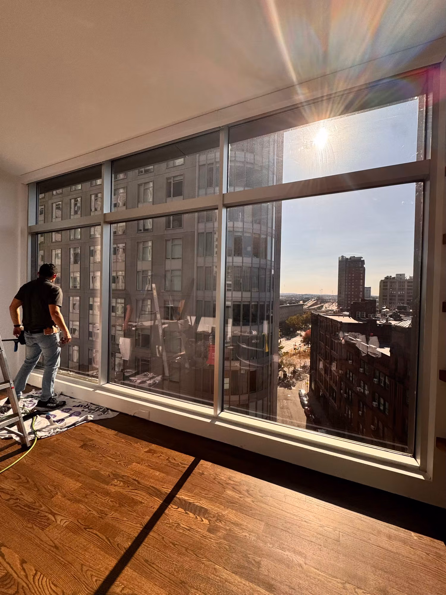 Technician installing solar control film on tall floor-to-ceiling windows with afternoon sun streaming through the glass.