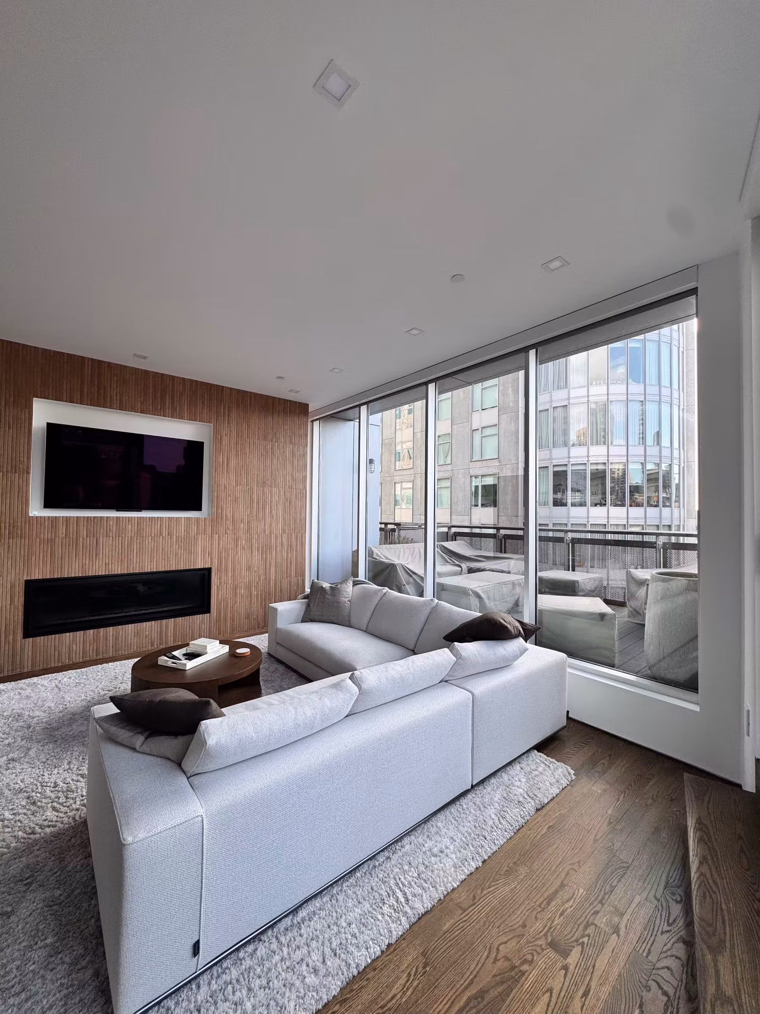 Modern living room with a fireplace, mounted television, and floor-to-ceiling corner windows facing the city.
