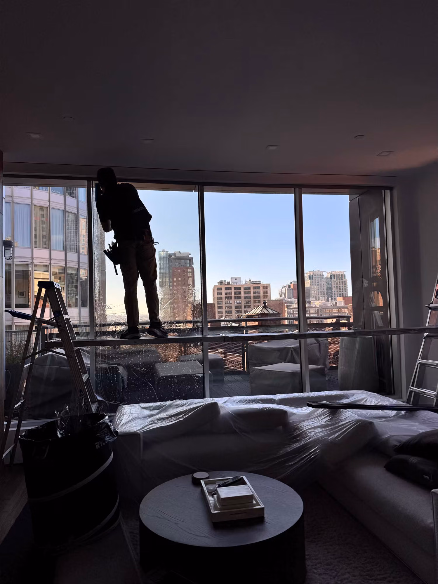 Installer applying solar control film to floor-to-ceiling living room windows above a covered sectional sofa.