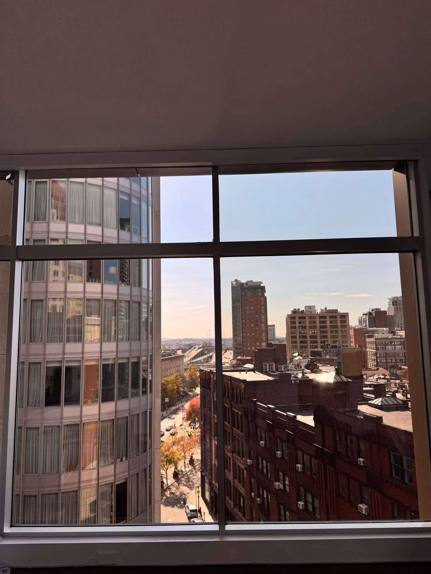 Large mullioned picture window overlooking a sunlit cityscape of high-rises, brick buildings, and a highway.