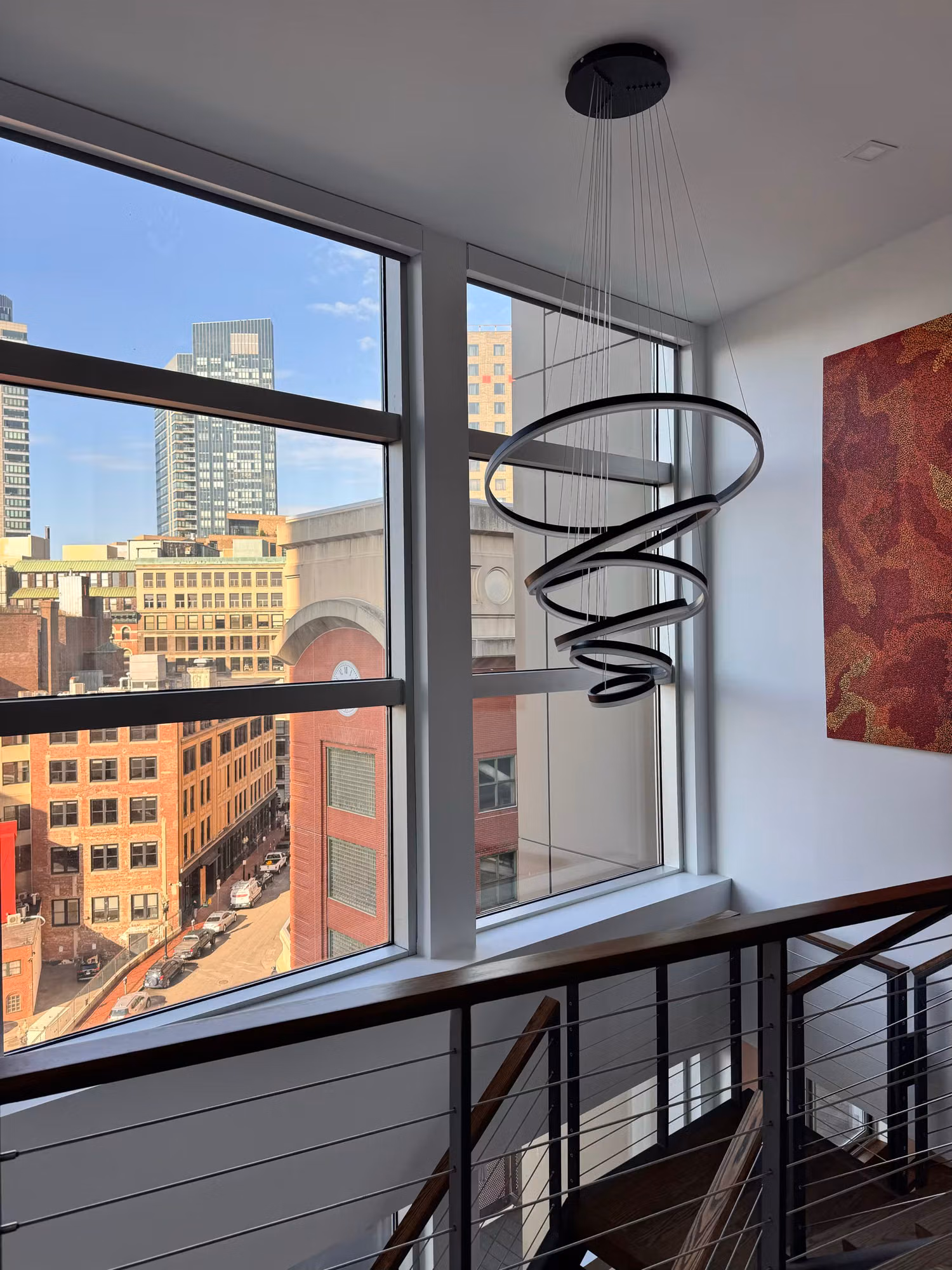 Tall stairwell picture window overlooking a city skyline above an open staircase with a sculptural pendant light.