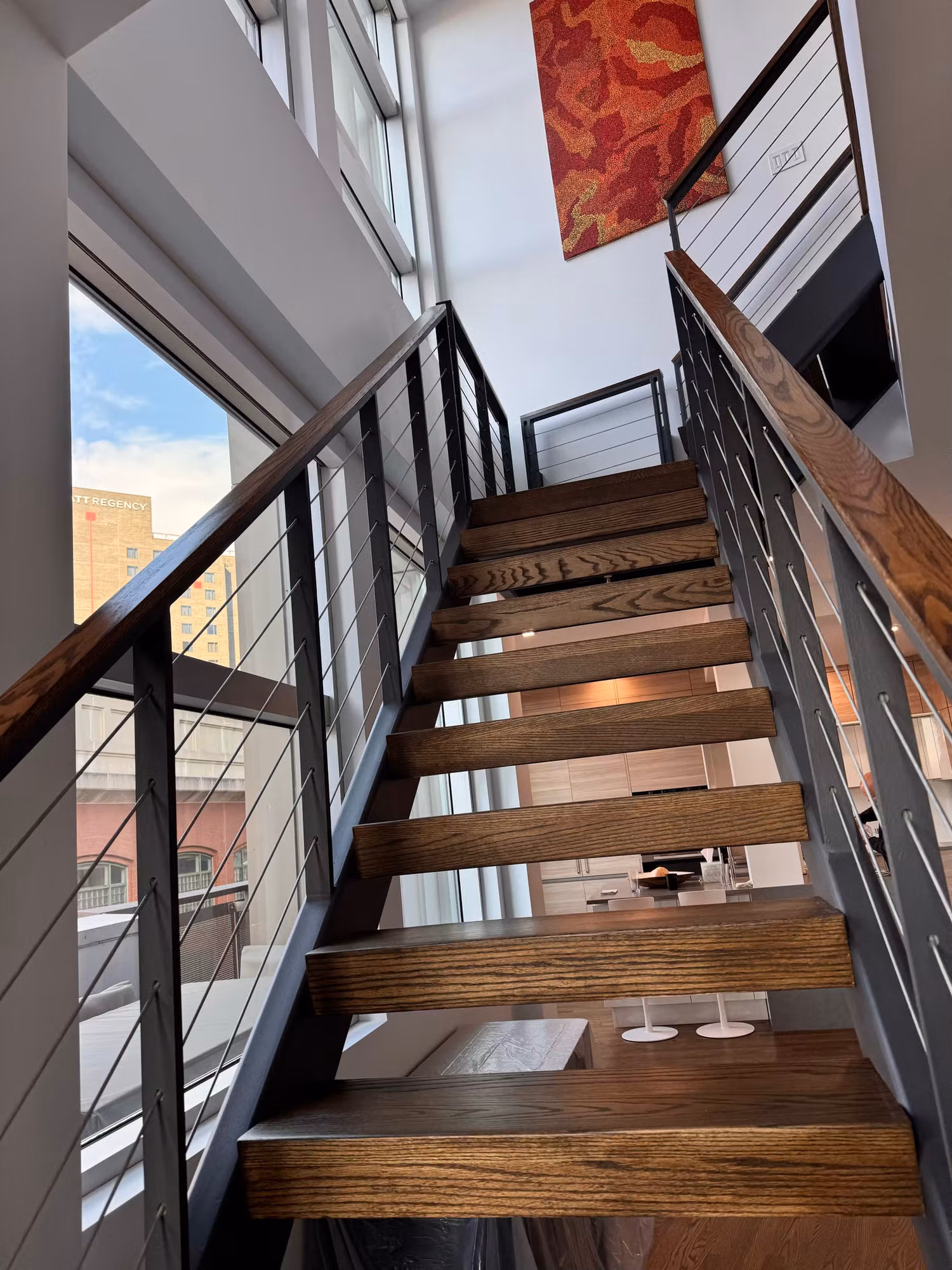 Open-tread wood staircase with cable railing beside a full-height window letting in daylight from the city.