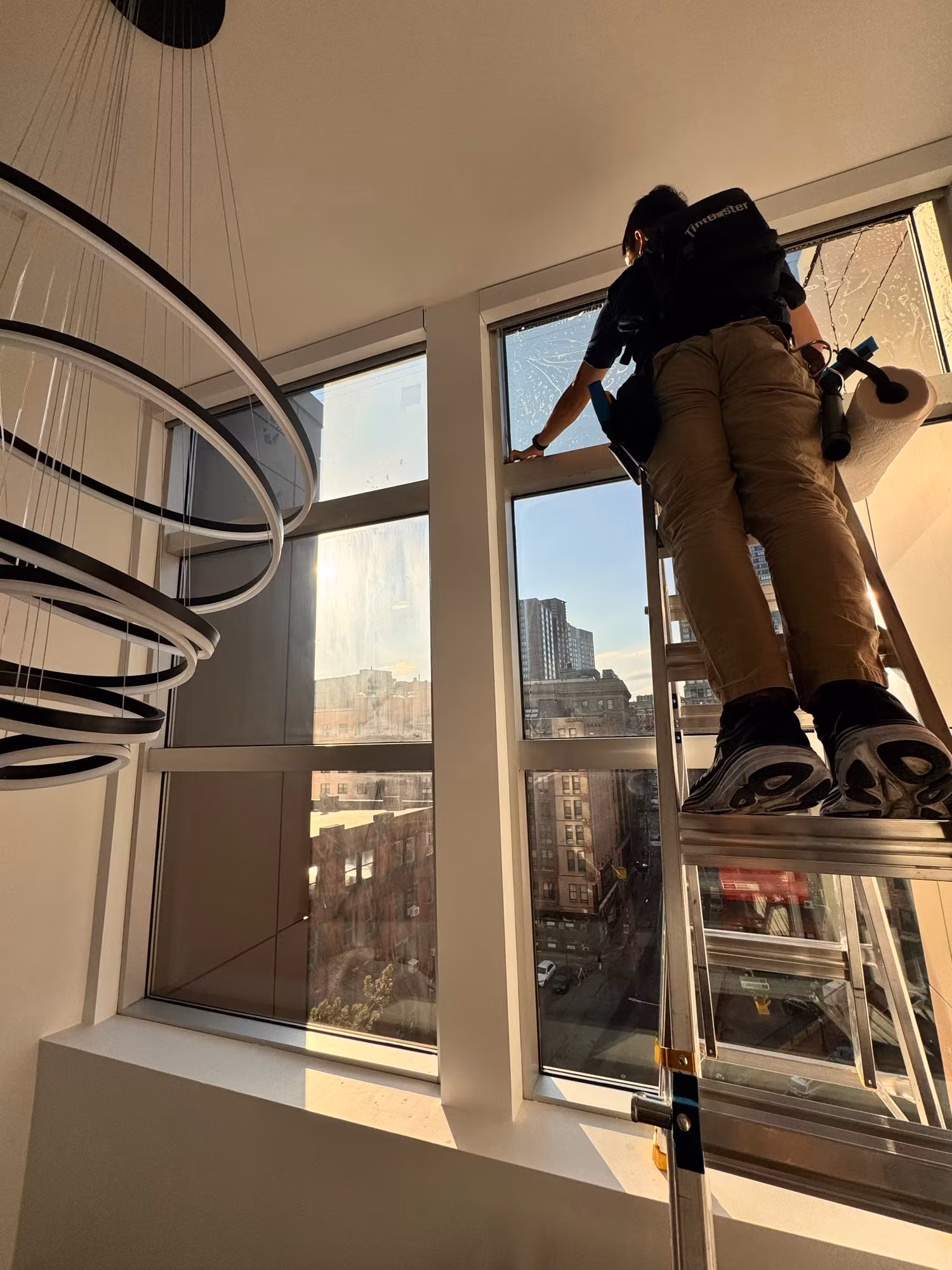 Technician on a ladder smoothing film onto a tall stairwell window near a ring chandelier and skyline.
