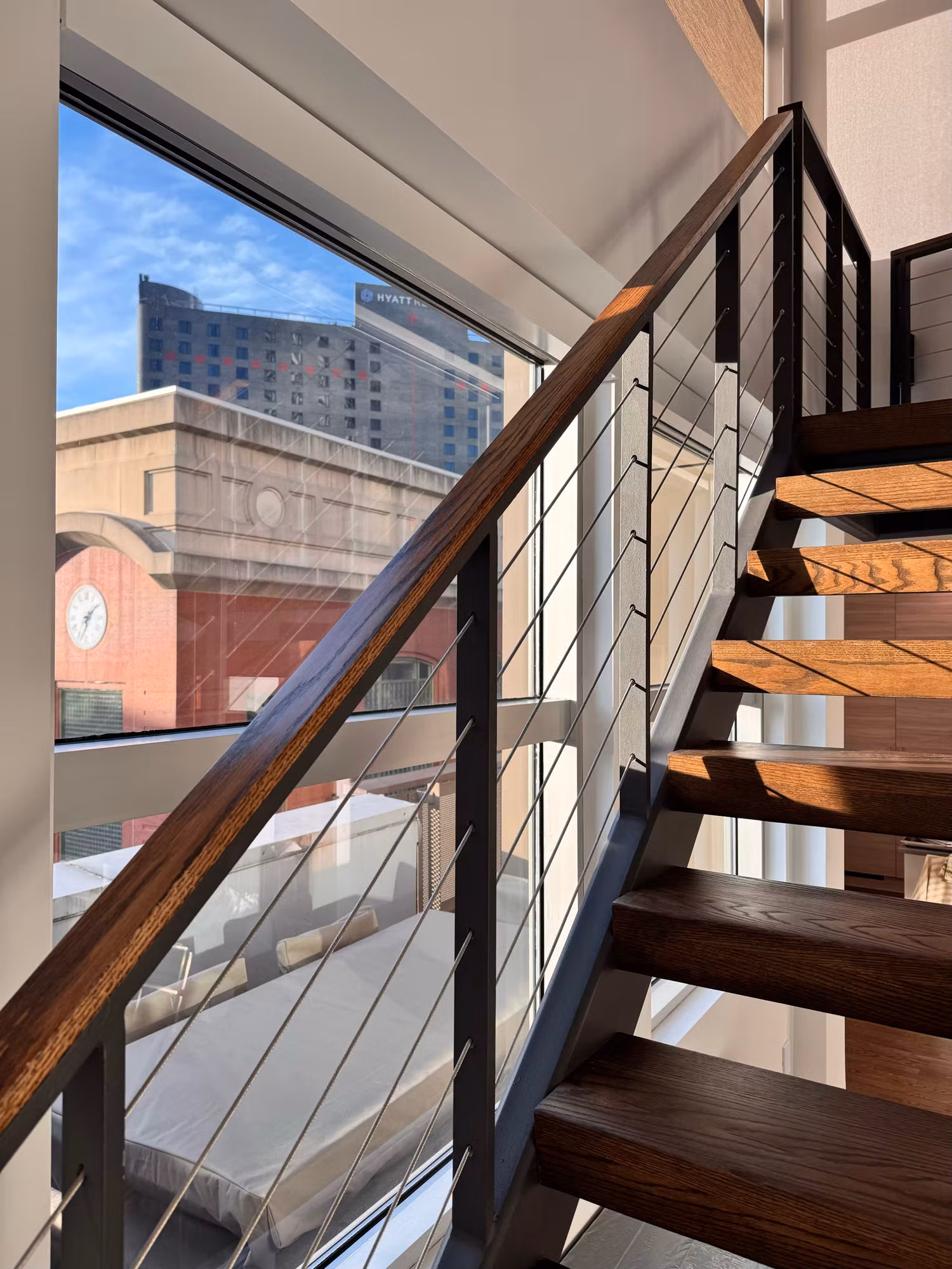 Stairwell window with solar control film overlooking nearby buildings beside a cable-rail wood staircase.