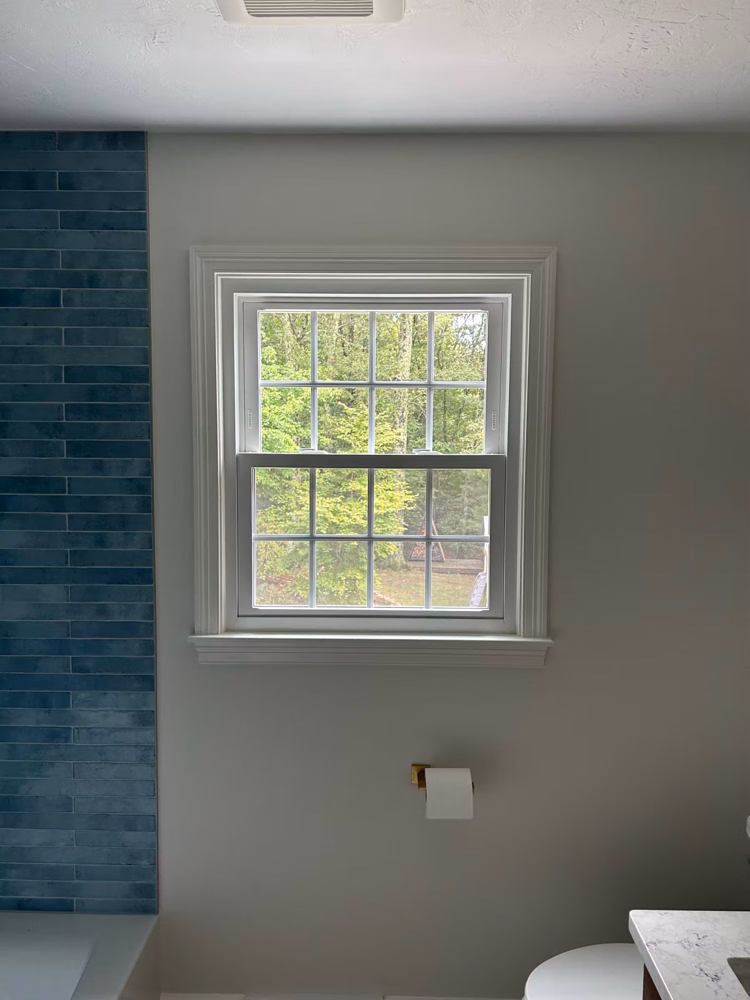 Bathroom window with white grilles beside a blue tile wall, looking out to trees, treated with clear safety film.