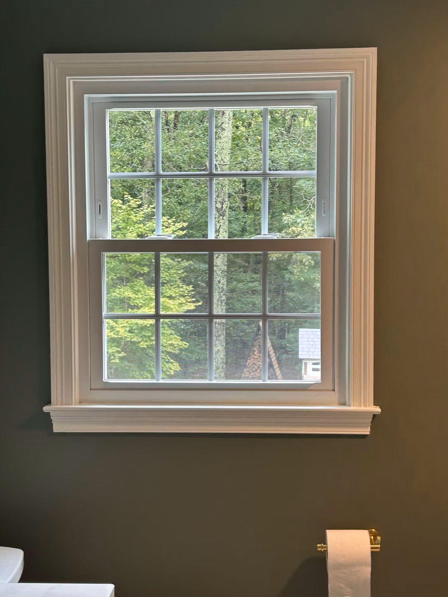 Bathroom double-hung window with white grilles on a dark wall, overlooking woods, fitted with clear safety film.