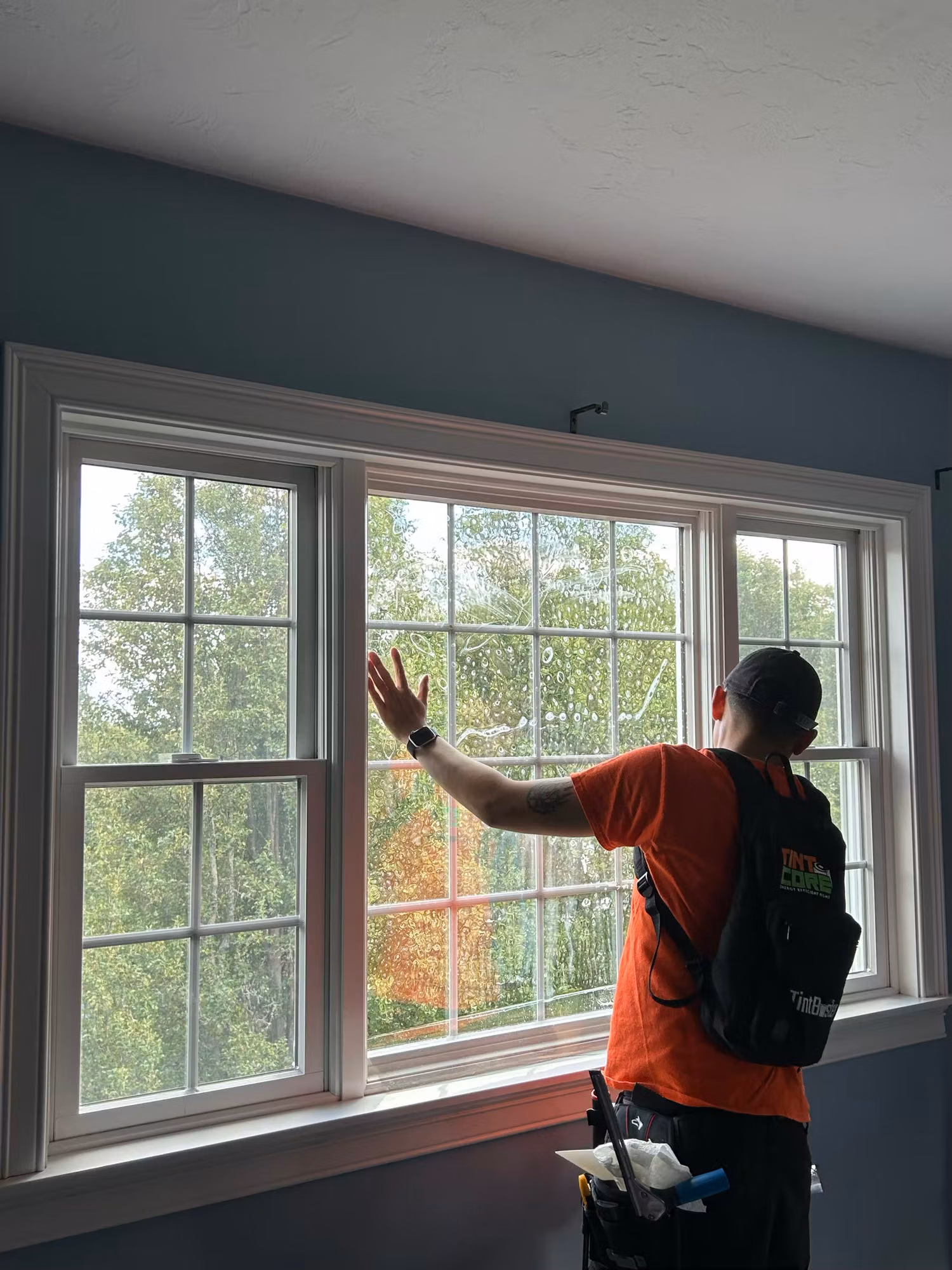 Installer applying clear window film to a triple bank of grid-pattern bedroom windows looking out onto autumn trees.