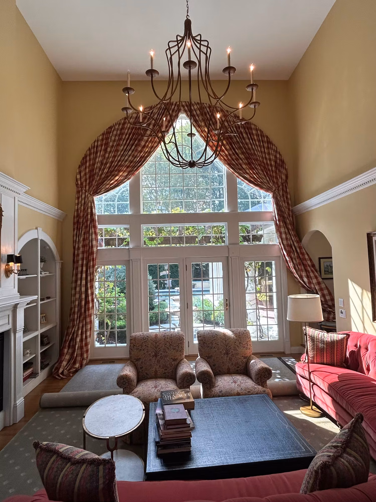 Tall arched window over French doors in a two-story living room with gingham drapes and chandelier.