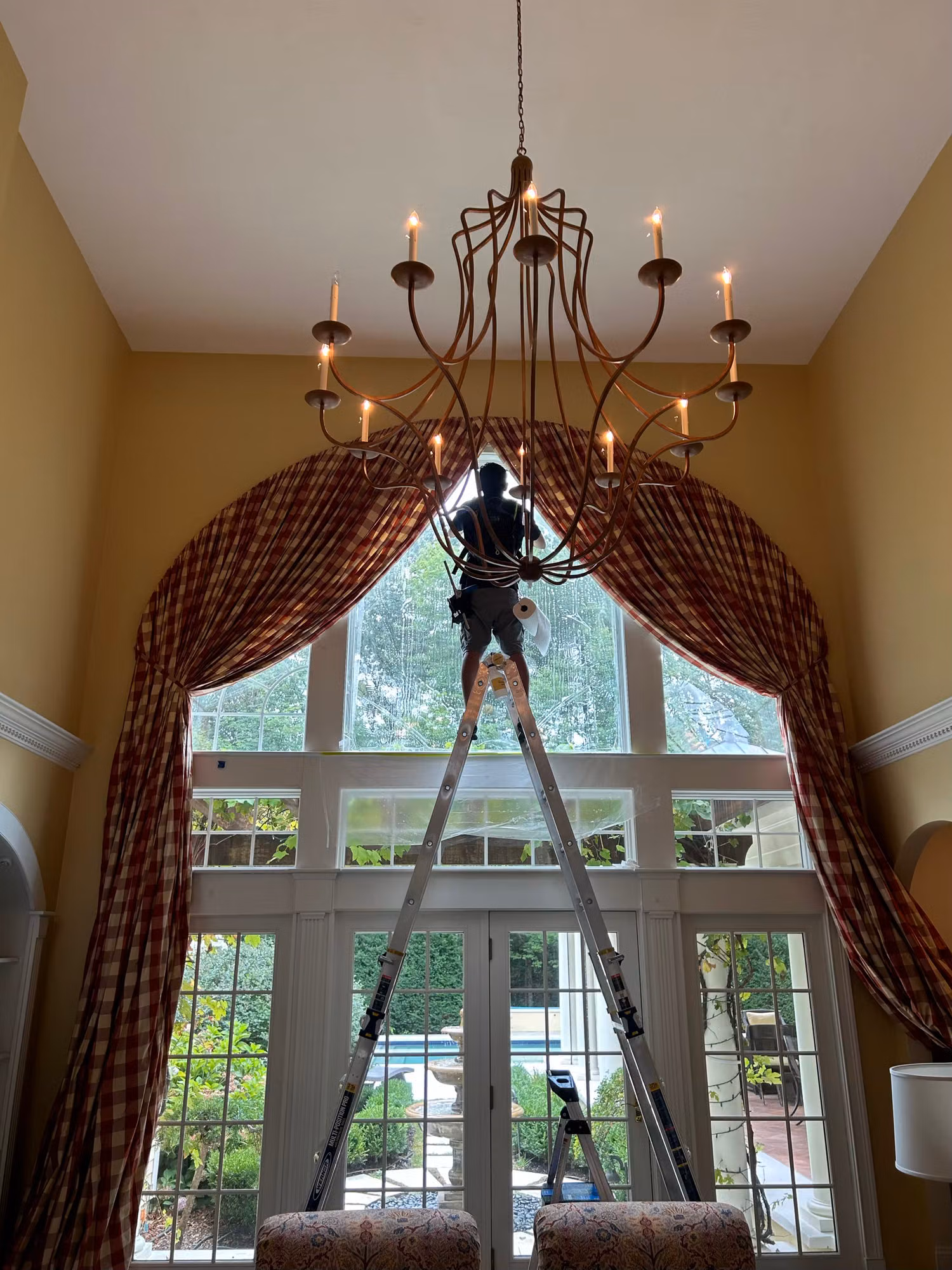 Installer on a tall ladder applying window film to a two-story arched window above French doors.