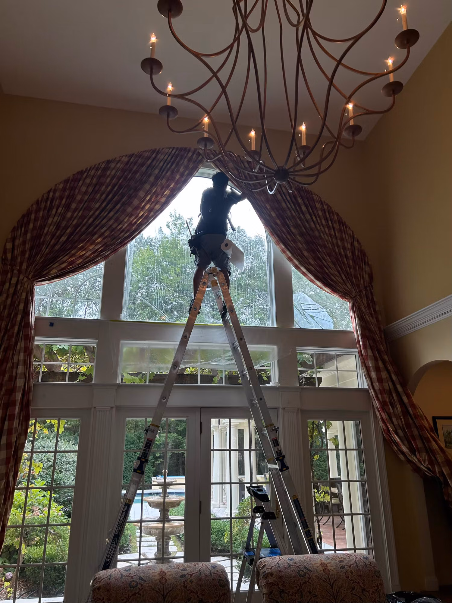Technician on a ladder installing film across the upper panels of a tall arched living room window.