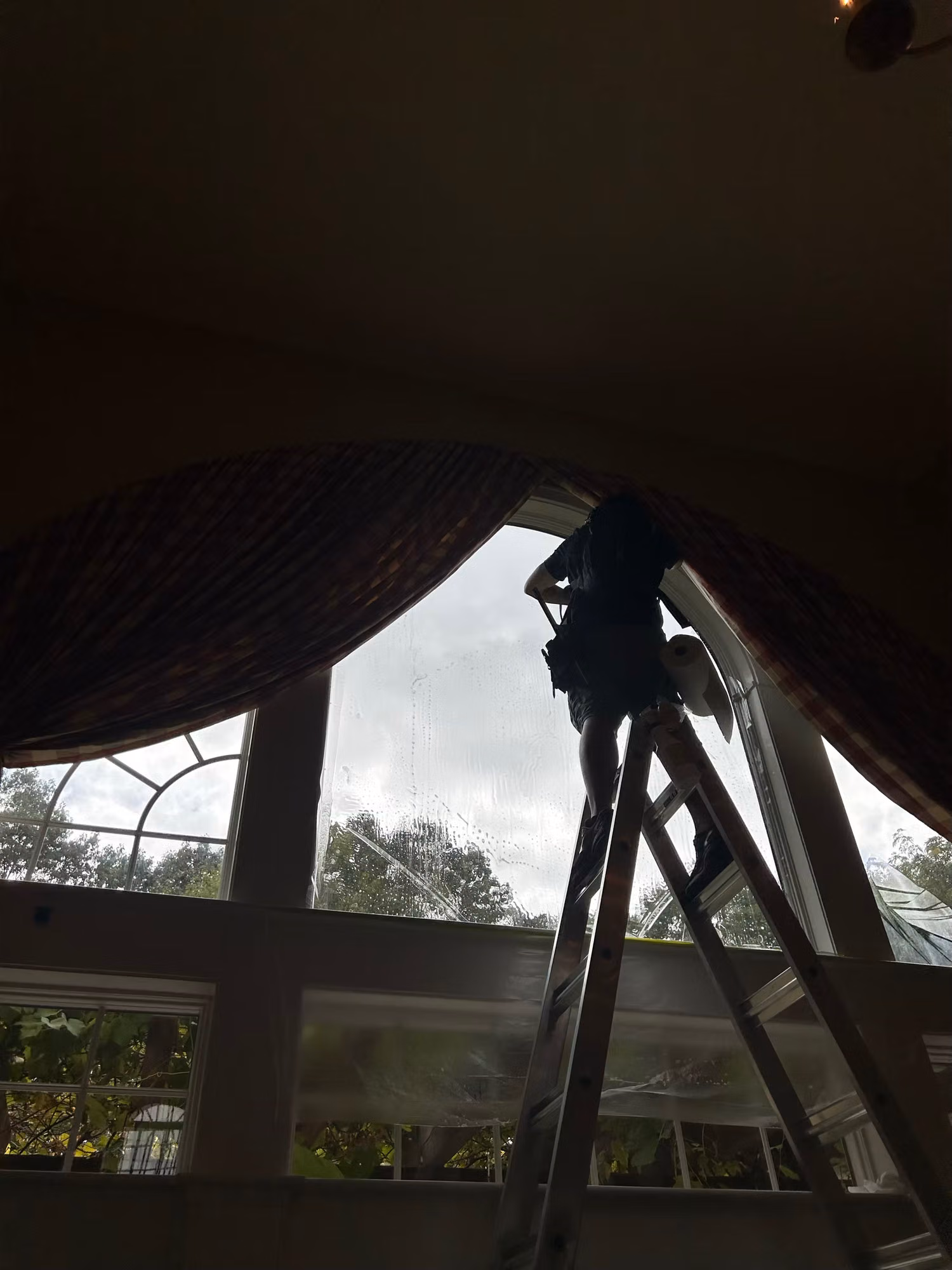 Backlit silhouette of an installer on a ladder smoothing film onto the upper glass of a tall window.
