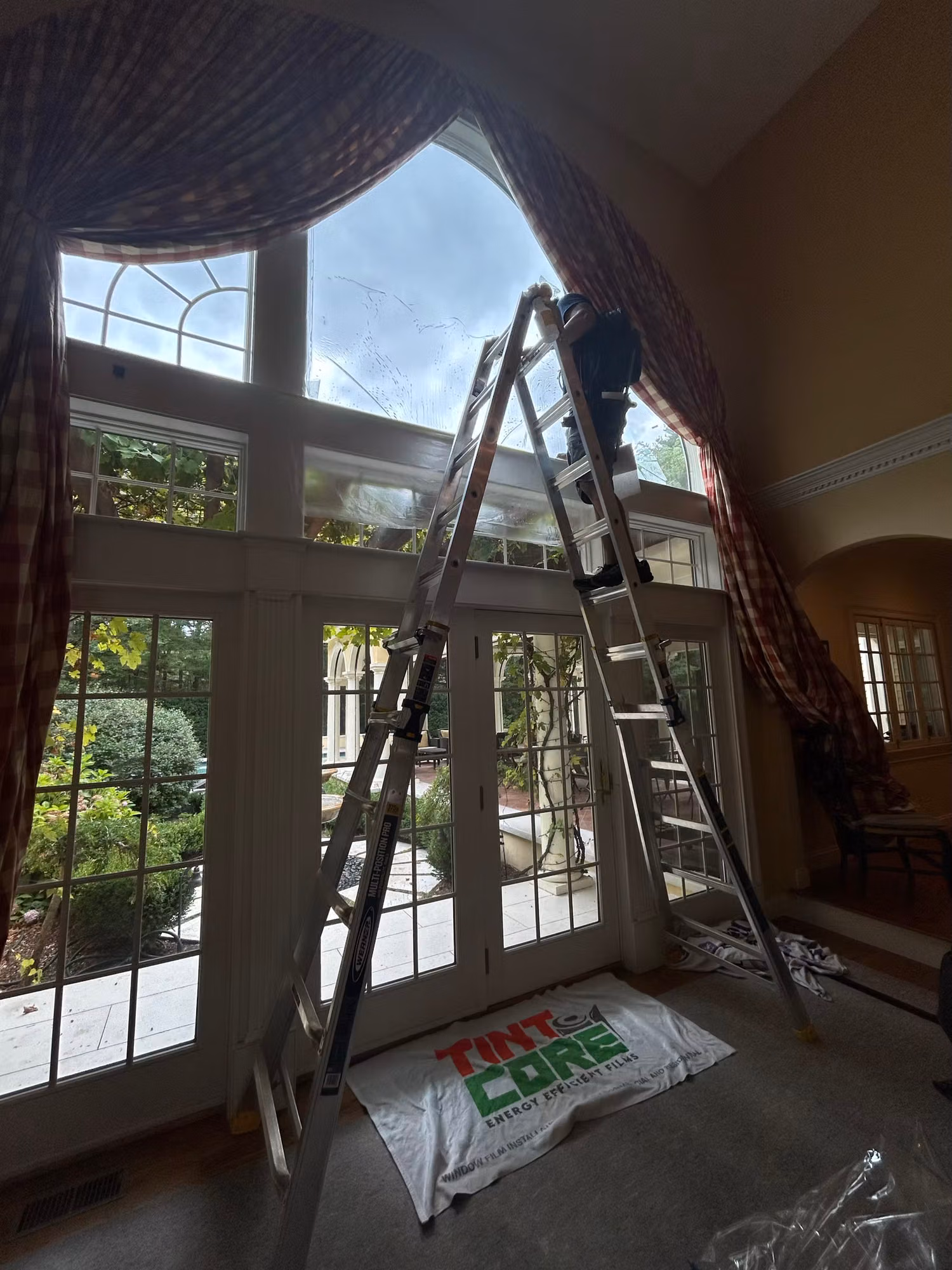 Installer on a ladder working film onto a two-story arched window above French doors in a living room.