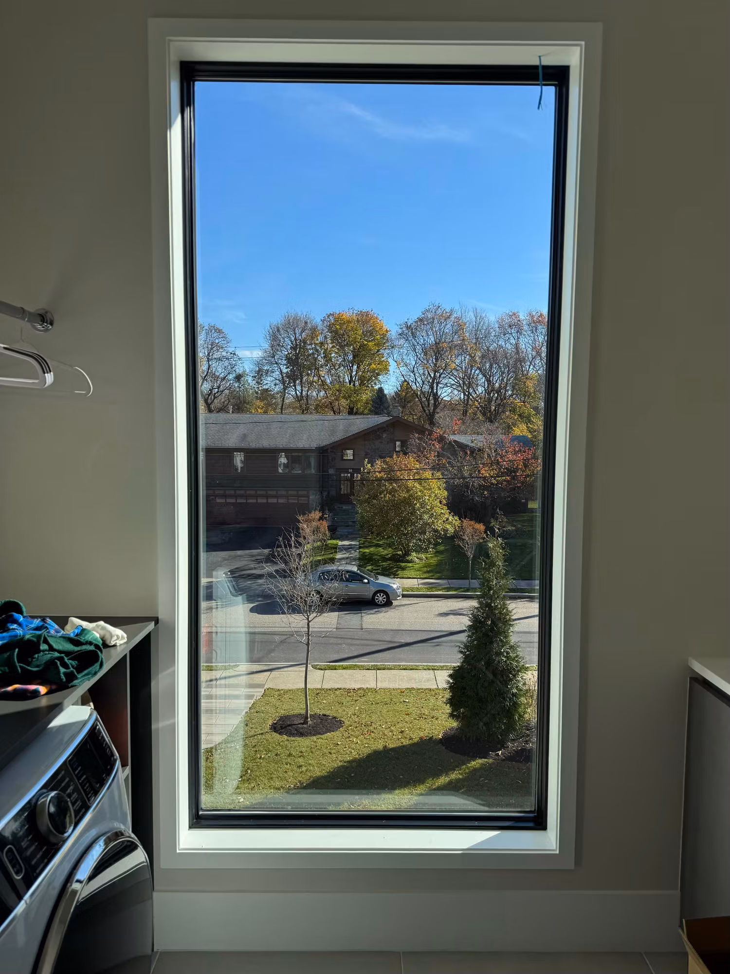 Tall laundry room window with clear glass before privacy film, looking out to a neighboring home and trees.