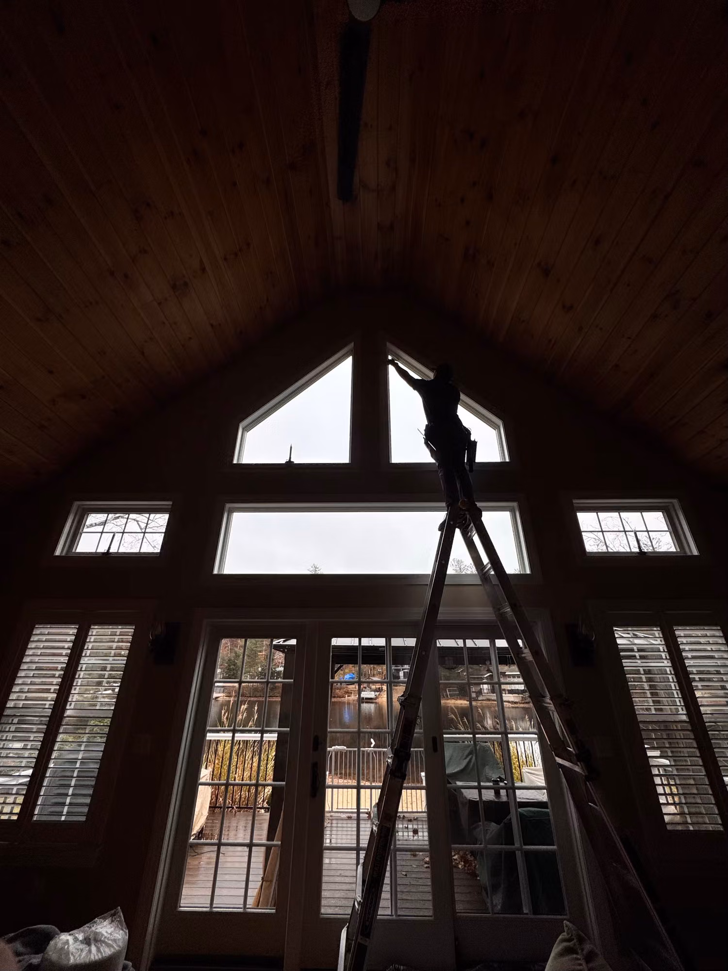 Installer on a ladder applying window film to the triangular peak window above a wall of transom windows and patio doors.