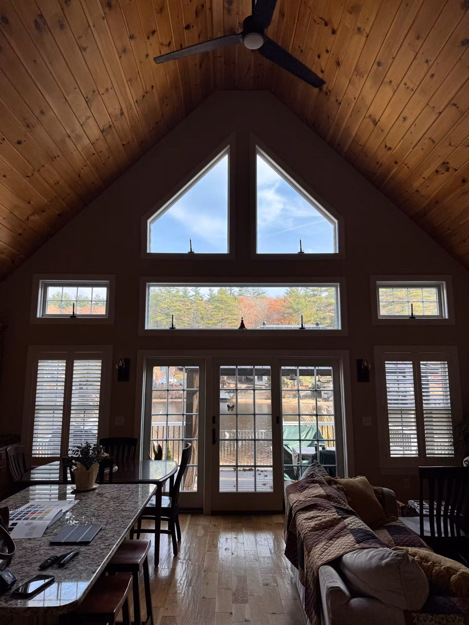 Vaulted great room with a tall window wall of triangular peak glass, transoms, and French patio doors framing a wooded view.