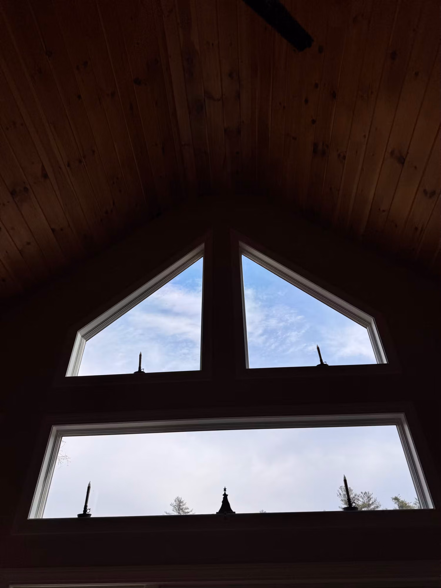 Triangular peak windows and a transom window set into a vaulted wood ceiling, glass looking out to a bright sky.