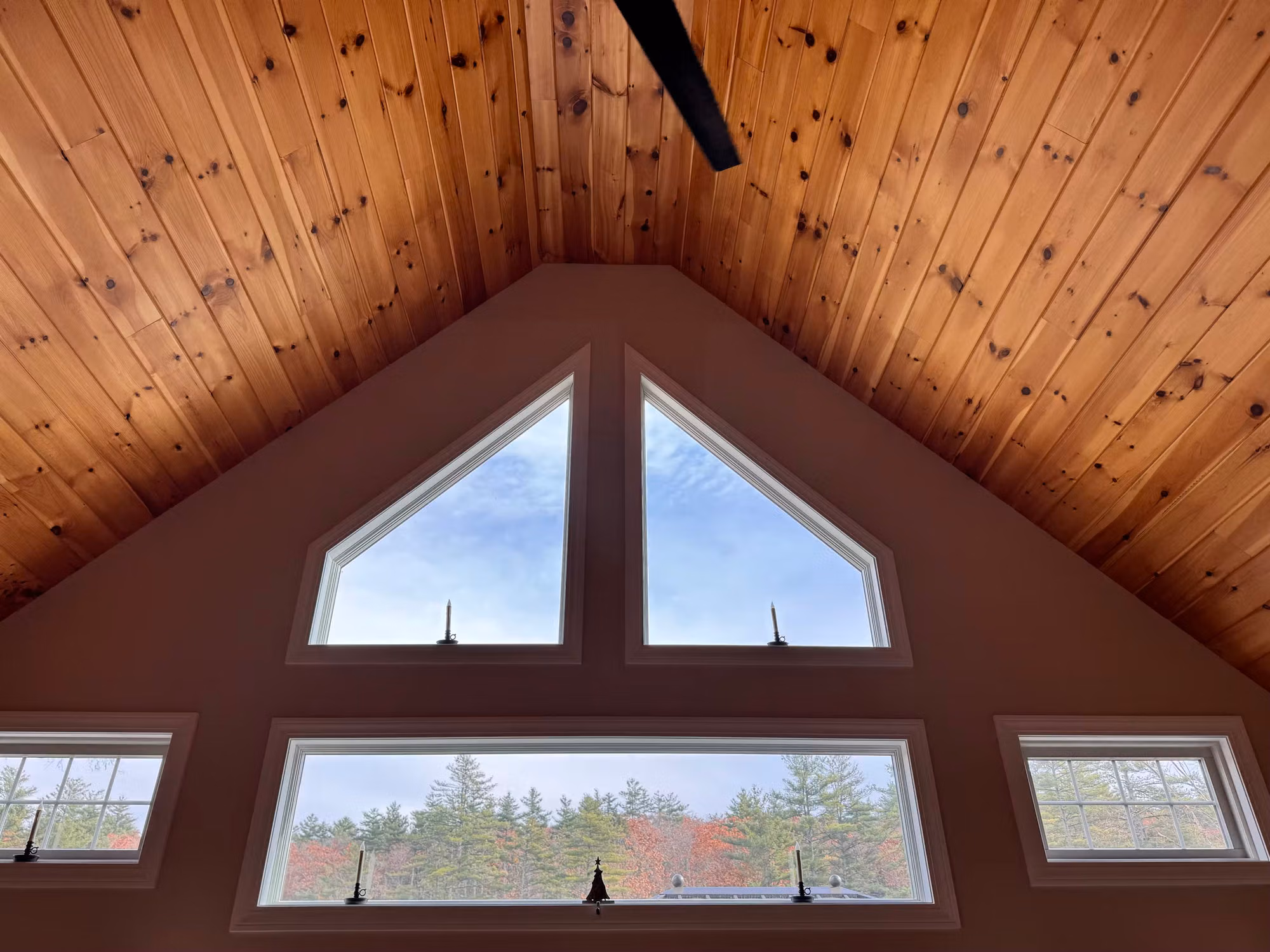 Twin triangular peak windows and lower transom windows under a knotty pine vaulted ceiling looking out to trees.