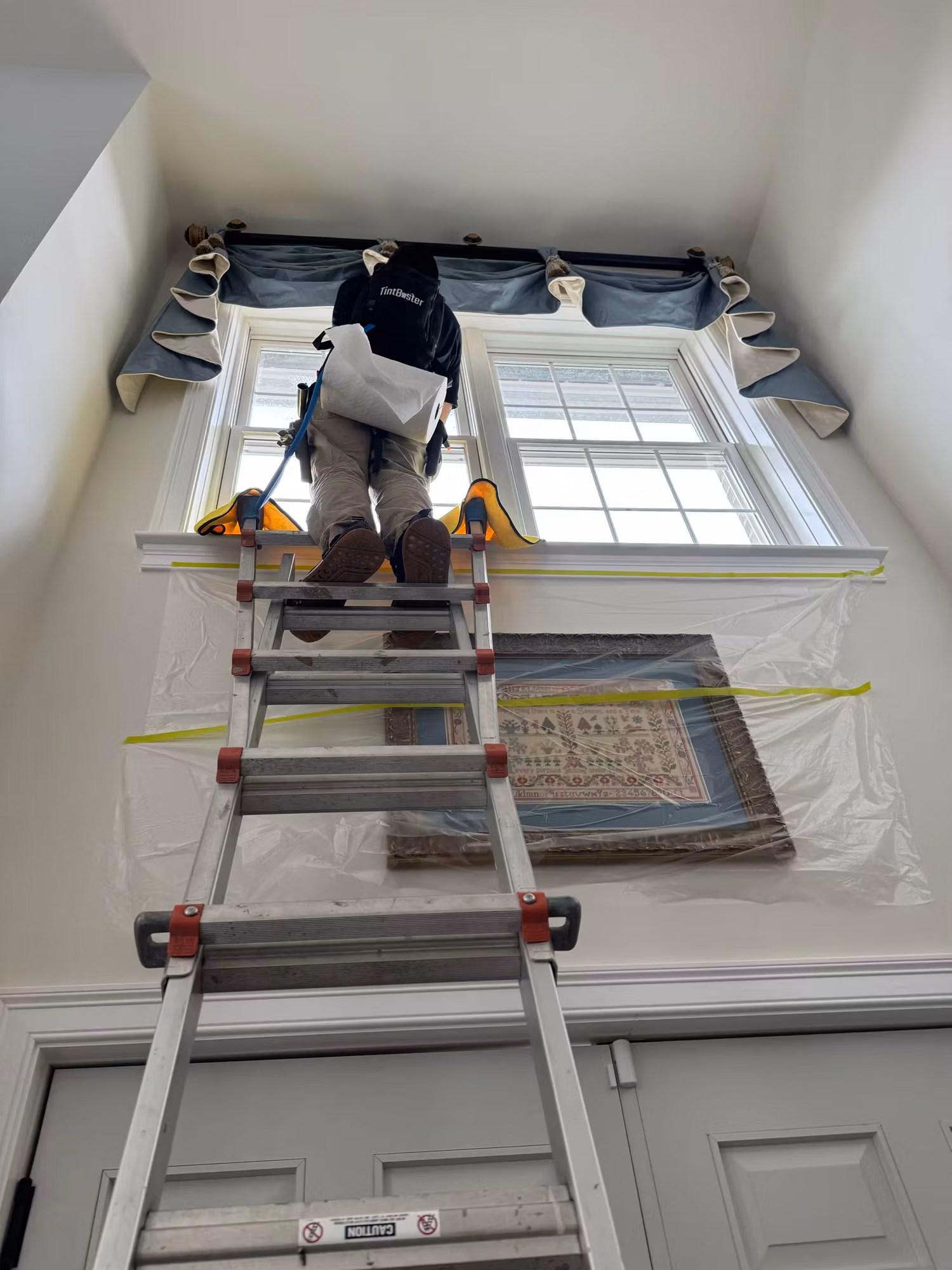 Installer on a ladder applying window film to a tall foyer window above an entry door, floor protected with plastic.